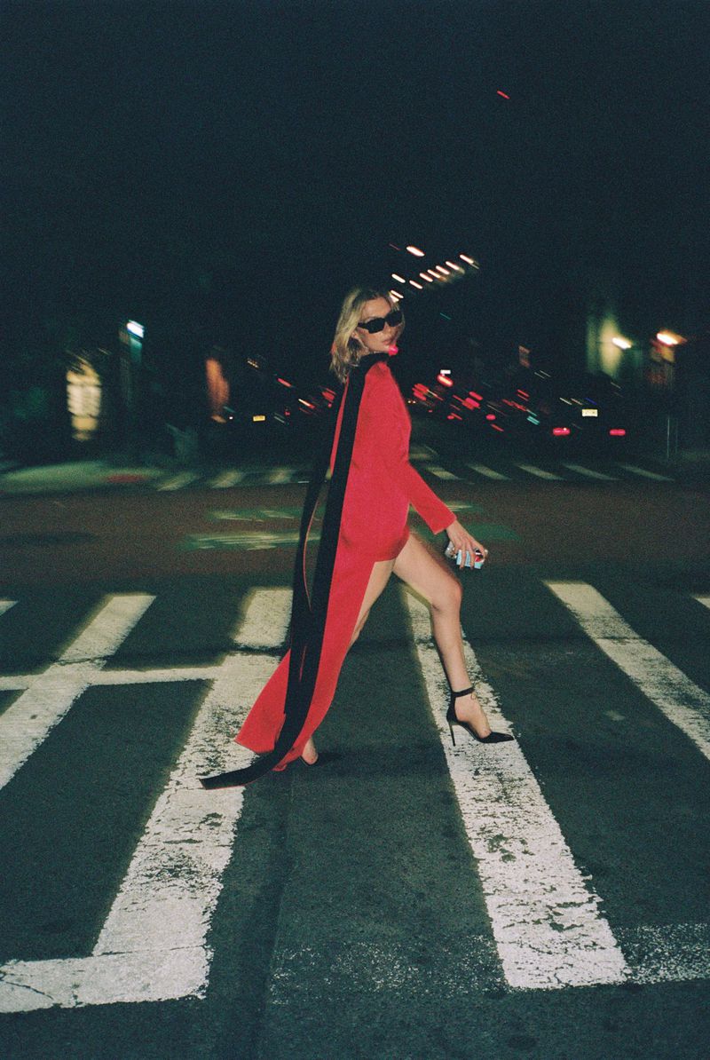 Woman in red dress with black cape walking across crosswalk at night. She's wearing sunglasses and black high heels in a city setting.