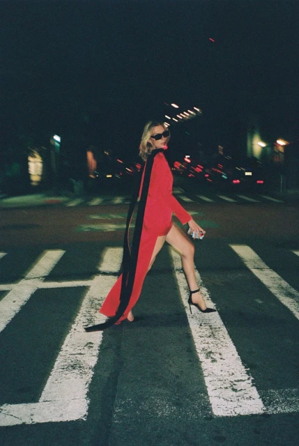 Woman in red dress with black cape walking across crosswalk at night. She's wearing sunglasses and black high heels in a city setting.