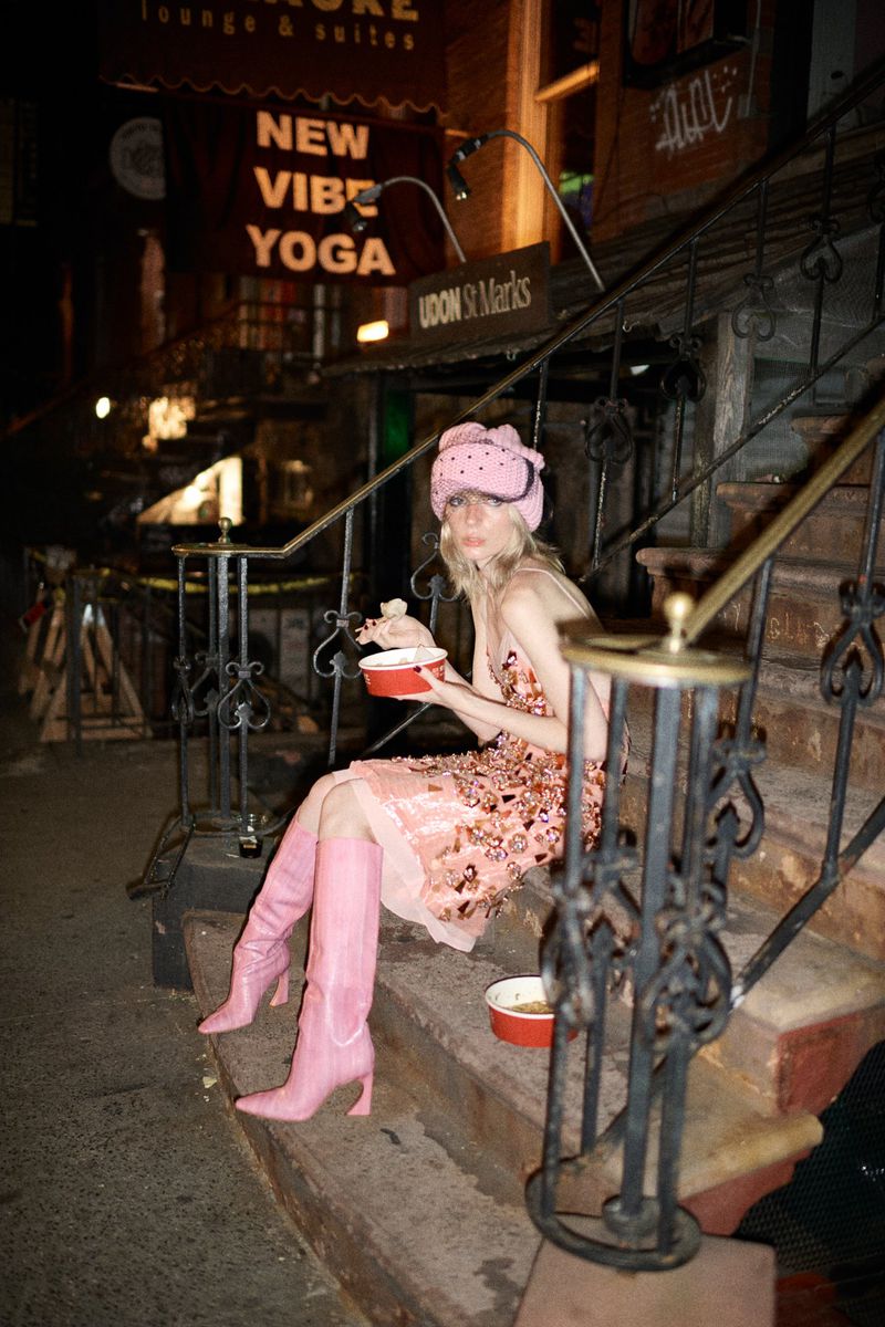 Woman in pink hat and boots sits on outdoor steps at night, holding bowl; restaurant sign visible in background with yoga advertisement.