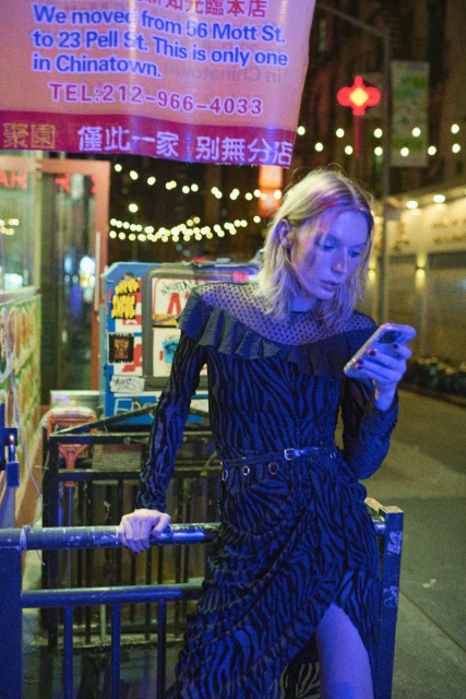 Woman in a patterned dress looks at her phone in a city street at night with buildings and signs visible in the background.