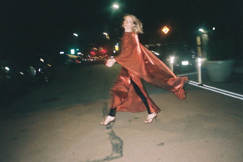 Woman in flowing red dress walking on city street at night. Streetlights illuminate the urban scene. She wears strappy heels.