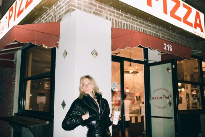 Blonde woman in a black leather jacket stands outside a pizza restaurant at night, holding a drink. Red awning above the restaurant entrance.