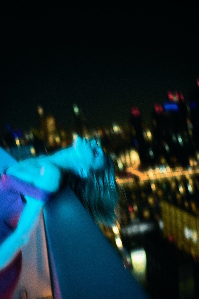 An adult female leans back on a rooftop ledge at night with city lights blurred in the background, lit with blue and violet tones.