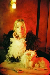 Model Willa Devereux wearing a black and white feather coat seated at a marble table under warm red lighting.