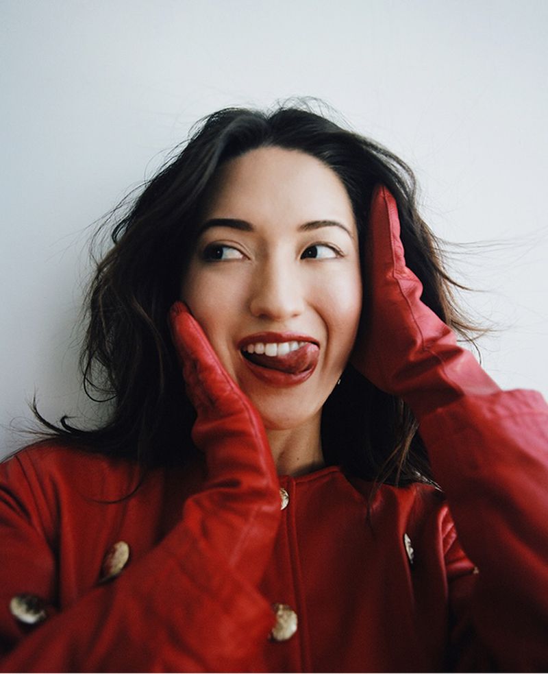 Portrait of a smiling woman with tongue sticking out, wearing red gloves and a red coat against a plain background, shot in natural light.
