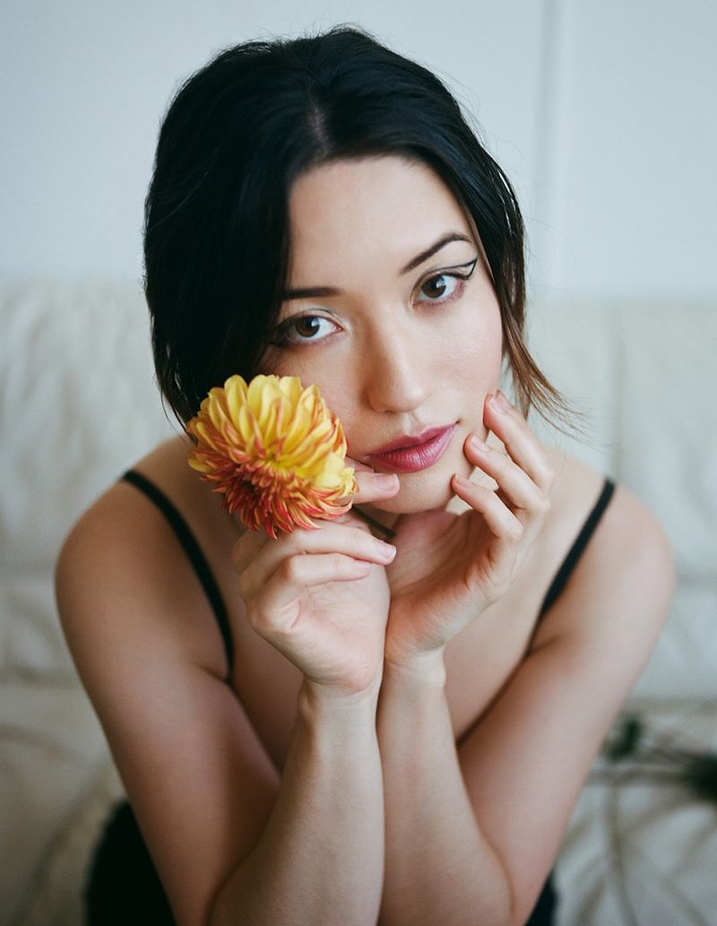 Attractive woman with dark hair holds a dahlia flower in a close-up portrait indoors with soft lighting and a simple, elegant background.