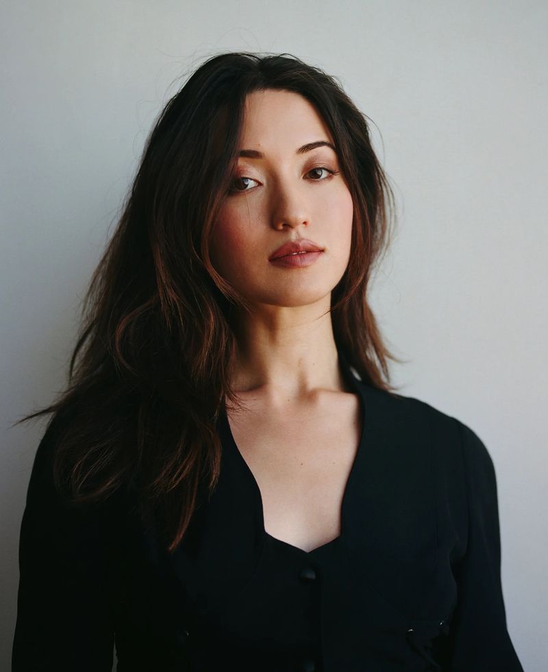 Portrait of a woman with long dark hair, wearing a black top, looking directly at the camera against a neutral background.