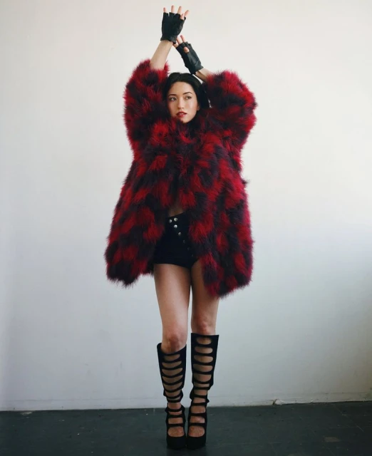A woman in black shorts, boots, and a red and black patterned coat poses with her hands up against a white background.