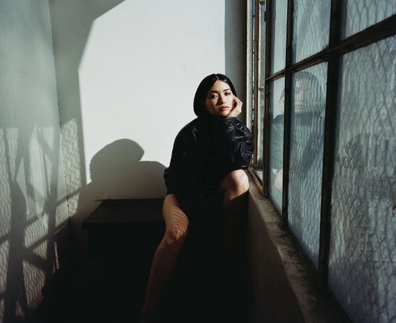 Young woman in oversized black jacket poses by a window with textured glass and shadow patterns. Seated, she looks toward the camera.