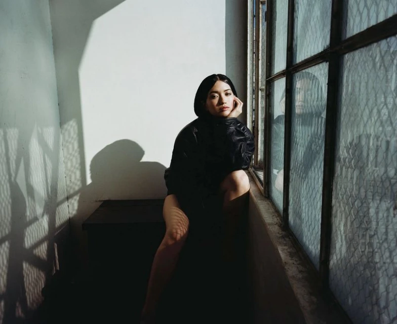 Young woman in oversized black jacket poses by a window with textured glass and shadow patterns. Seated, she looks toward the camera.
