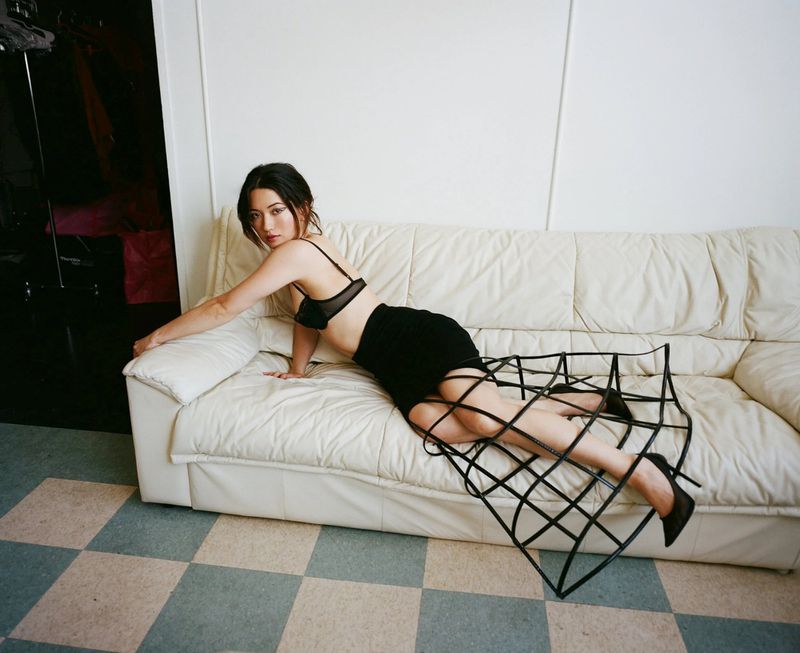 Young woman in bra and skirt lying on white couch with black metal sculpture. Indoor studio shot with checkered floor.