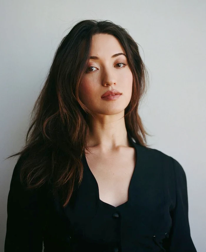 Portrait of Reina Hardesty wearing a black top with a notched neckline against a neutral pale grey studio background.