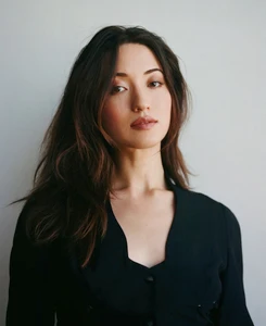 Portrait of Reina Hardesty wearing a black top with a notched neckline against a neutral pale grey studio background.
