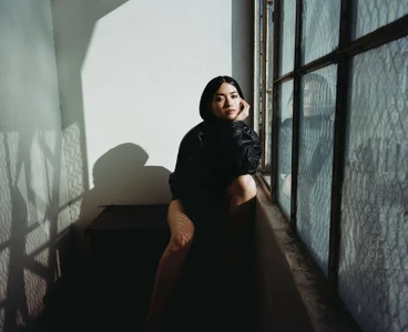 Portrait of Reina Hardesty seated by a grid-patterned window wearing a dark jacket for Numero Netherlands by Lauren Withrow