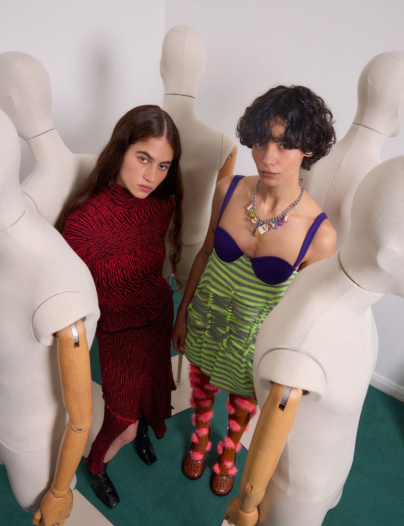 Two fashion models pose with mannequin figures in a studio setting, showcasing bold and avant-garde clothing and accessories.