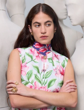 Woman in floral dress with gold jewelry posing in front of neutral-toned mannequins, sleeveless, pink blossoms, portrait framing.