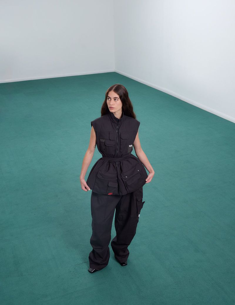 A woman stands in a bright room with blue-green flooring, modeling a black sleeveless vest and a voluminous black skirt over black pants.