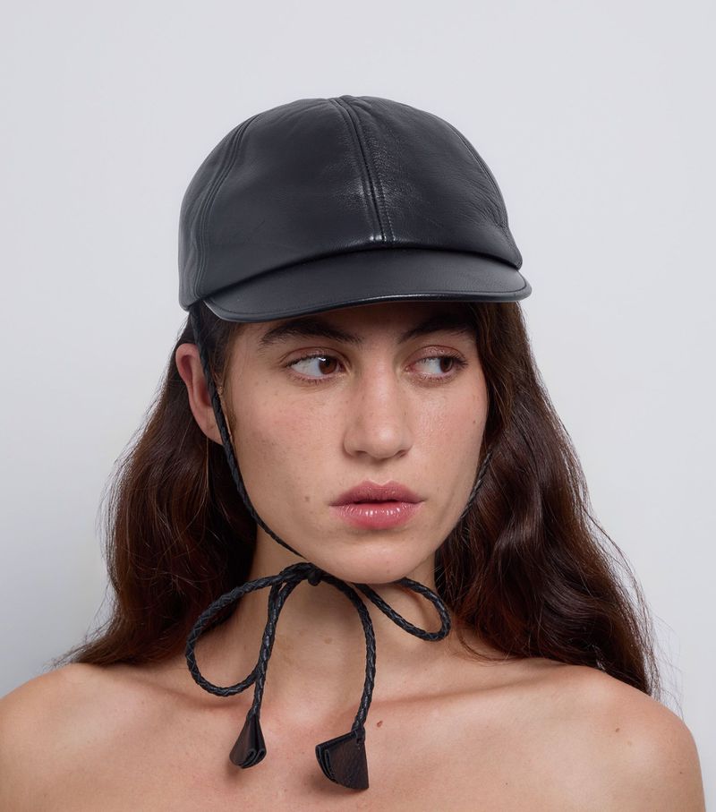 Close-up of a woman with long, dark hair wearing a black leather hat with a tied braided leather string beneath her chin against white.