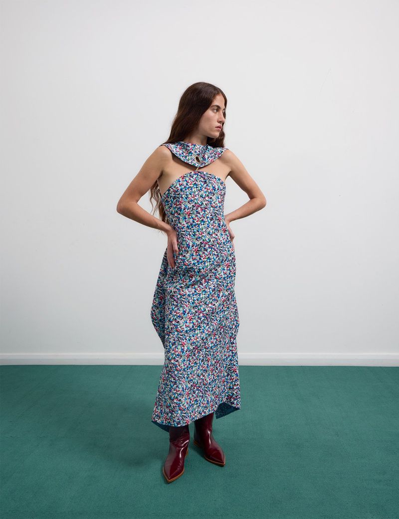An adult woman with long brown hair poses in a colorful floral dress and shiny red boots against a white and green backdrop.