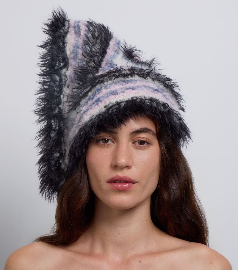 Close-up of a woman with long brown hair wearing a furry striped hat against a pale grey background in a studio fashion shot.