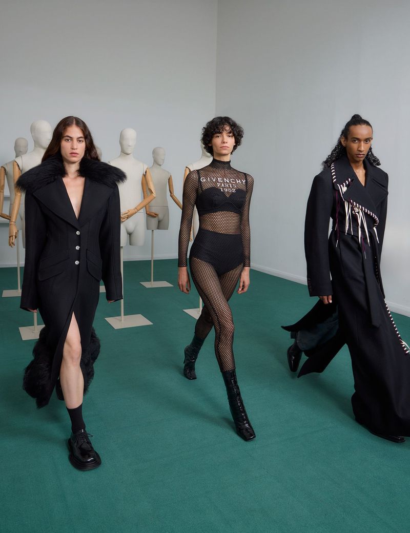 Three fashion models in black outfits walk towards the camera in a minimalist studio with mannequin torsos in the background.