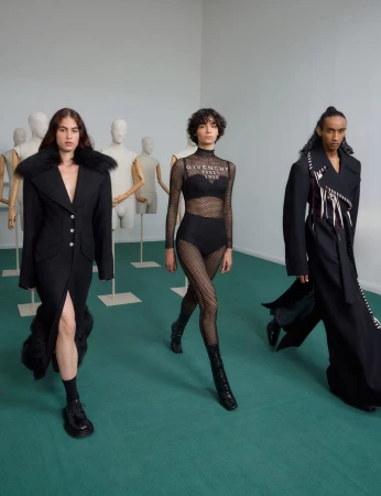 Three fashion models in black outfits walk towards the camera in a minimalist studio with mannequin torsos in the background.