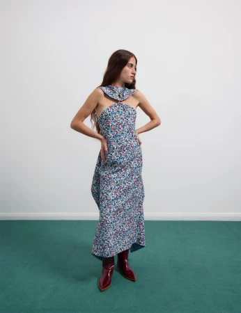 Model Iris O'Carroll poses in a floral print halter dress and glossy burgundy boots on a teal carpet against a white wall.