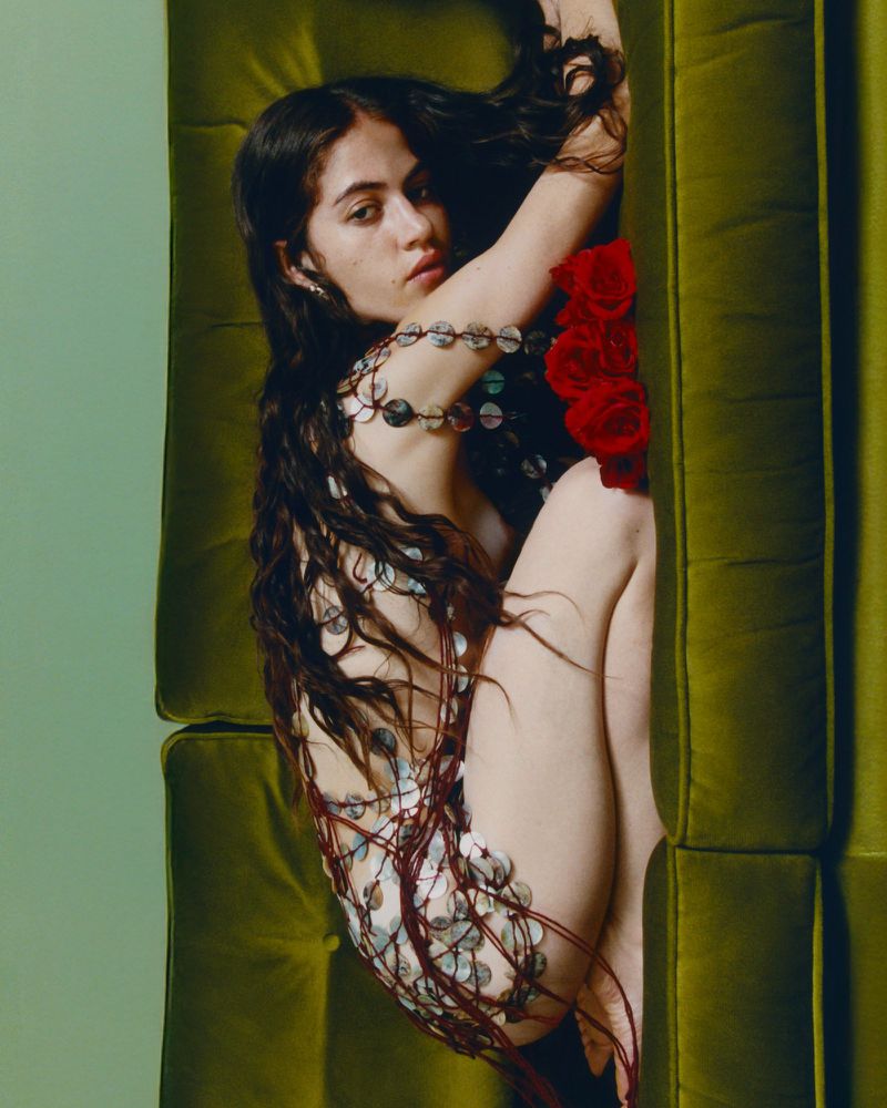 Young woman with long dark hair reclines on green cushion wearing beaded top and holding red roses, looking at the camera.