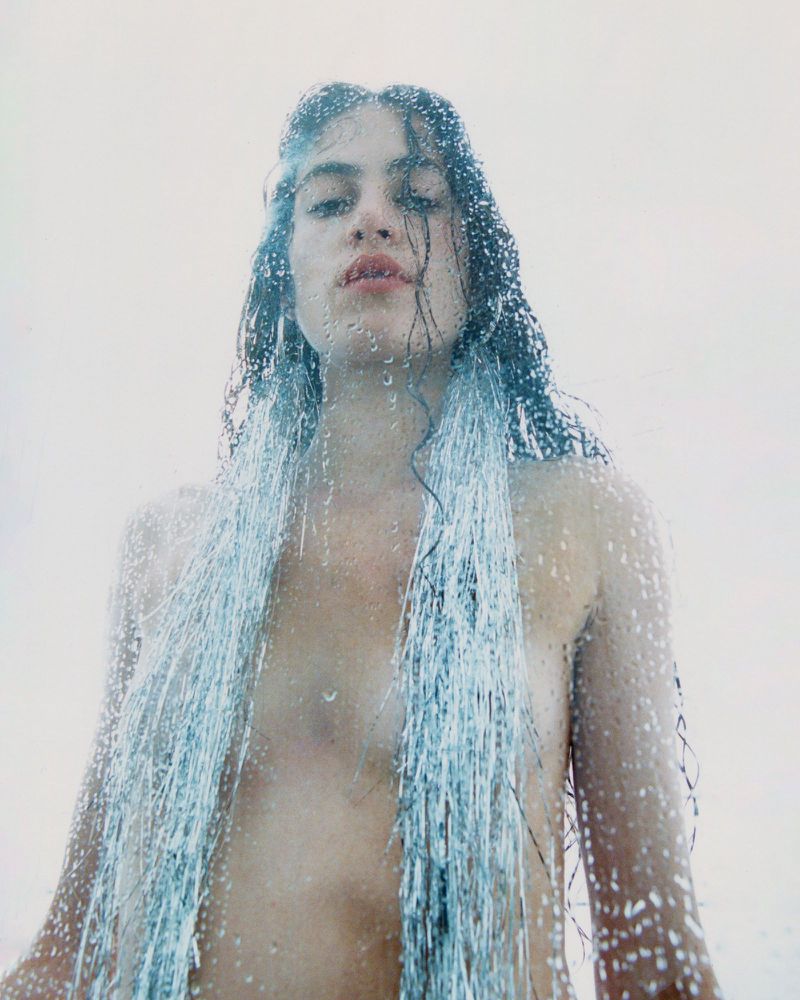 Woman with wet hair and tinsel standing behind a wet glass surface, creating a blurred and intimate close-up studio portrait.