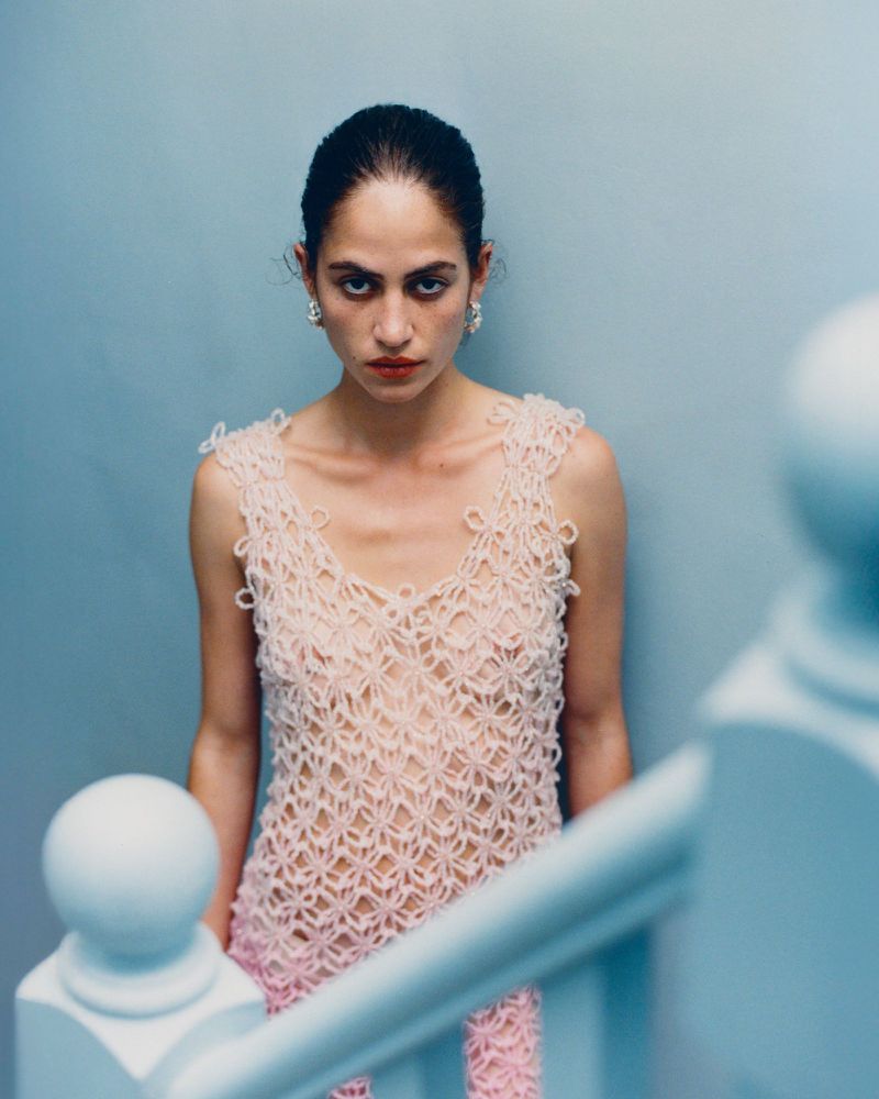 Model Iris O'Carroll wears a pale pink crochet open-knit dress, standing by a blue staircase banister against a pale blue wall.