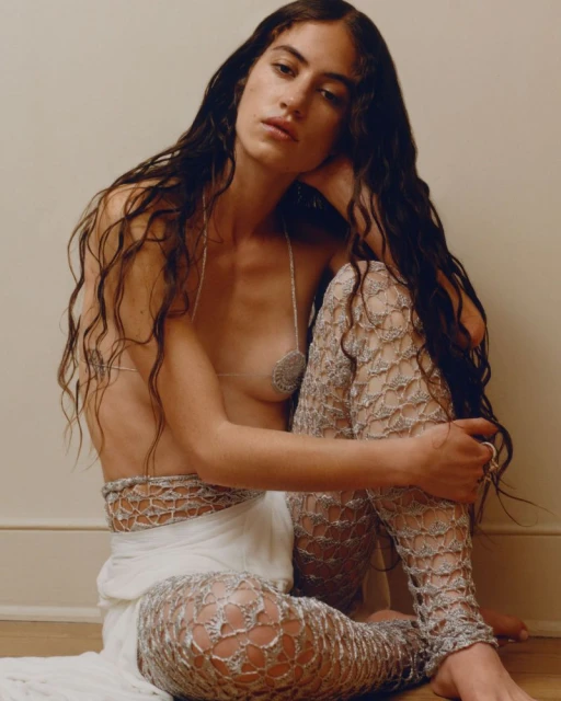 Portrait of an adult female with long, dark wavy hair posing indoors in a silver top and leggings against a neutral wall.