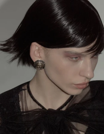 Close-up of a female model with dark hair and an earring, wearing a black lace top, against a neutral grey backdrop in a photo studio.