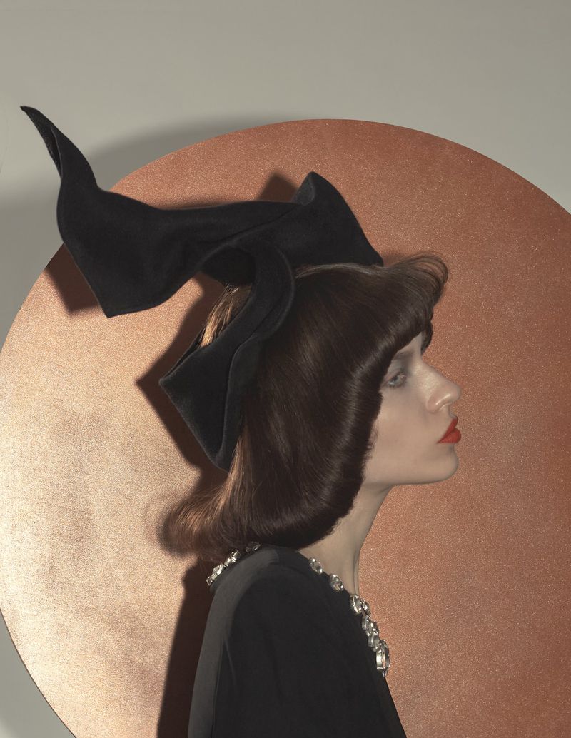 Woman with dark hair styled with a large black bow, wearing a black dress and jeweled necklace, set against a textured, circular backdrop.