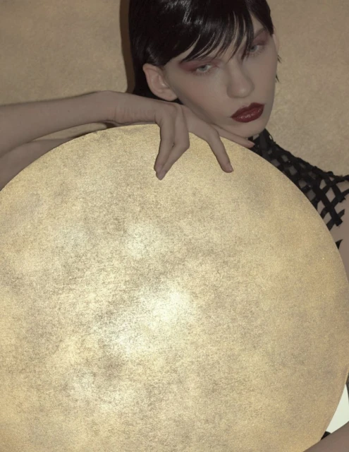 A young woman with dark hair and red lipstick poses with her hand resting on a large, glowing, circular lamp against a textured background.
