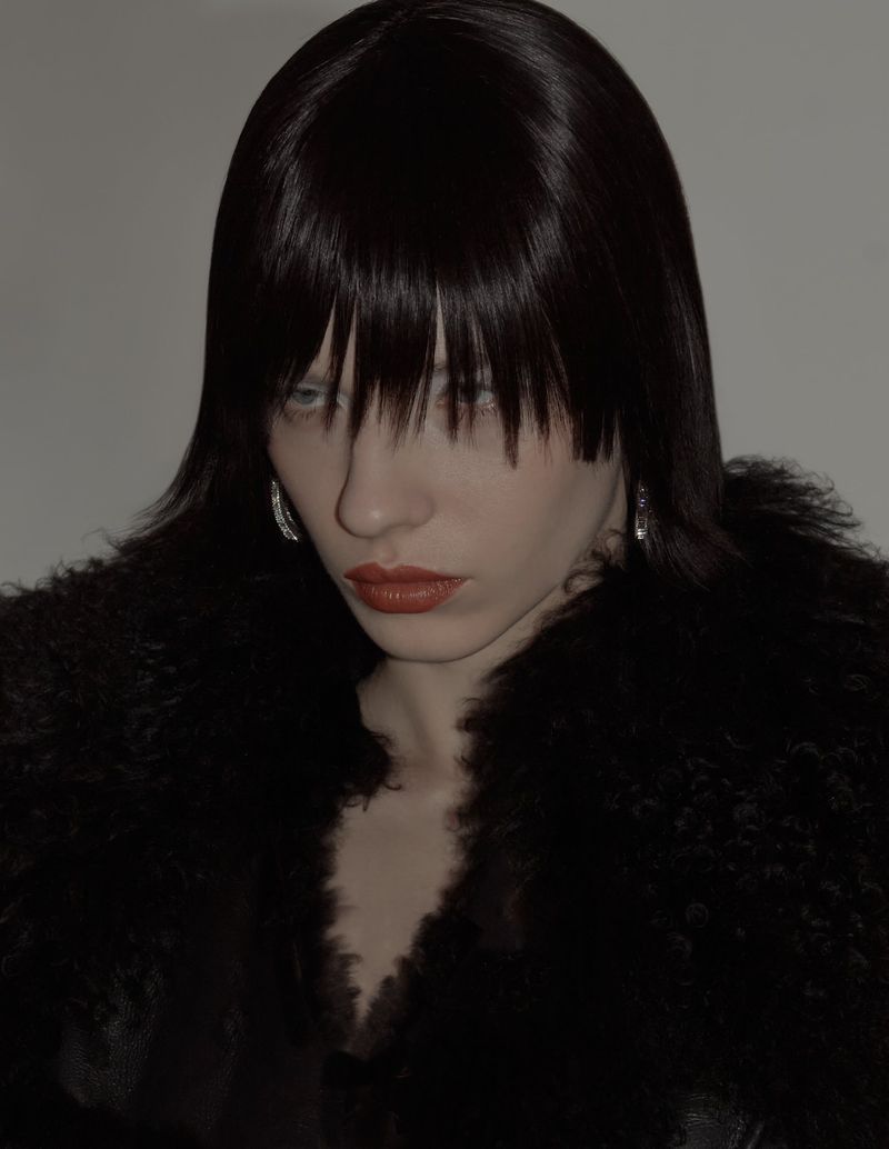 Kate Letskaya with straight dark bangs and red lipstick wearing a black textured fur collar against a grey background.