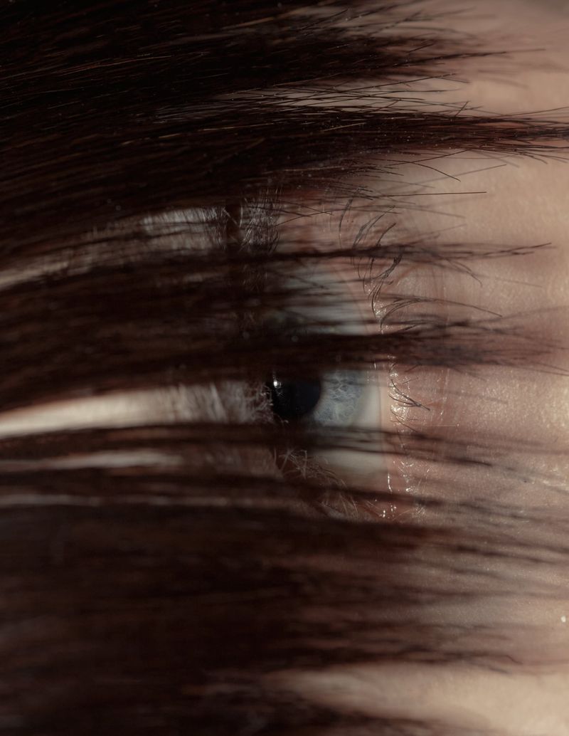 Close-up of a person's eye visible through strands of dark brown hair that partially obscure the view, focusing on the iris.