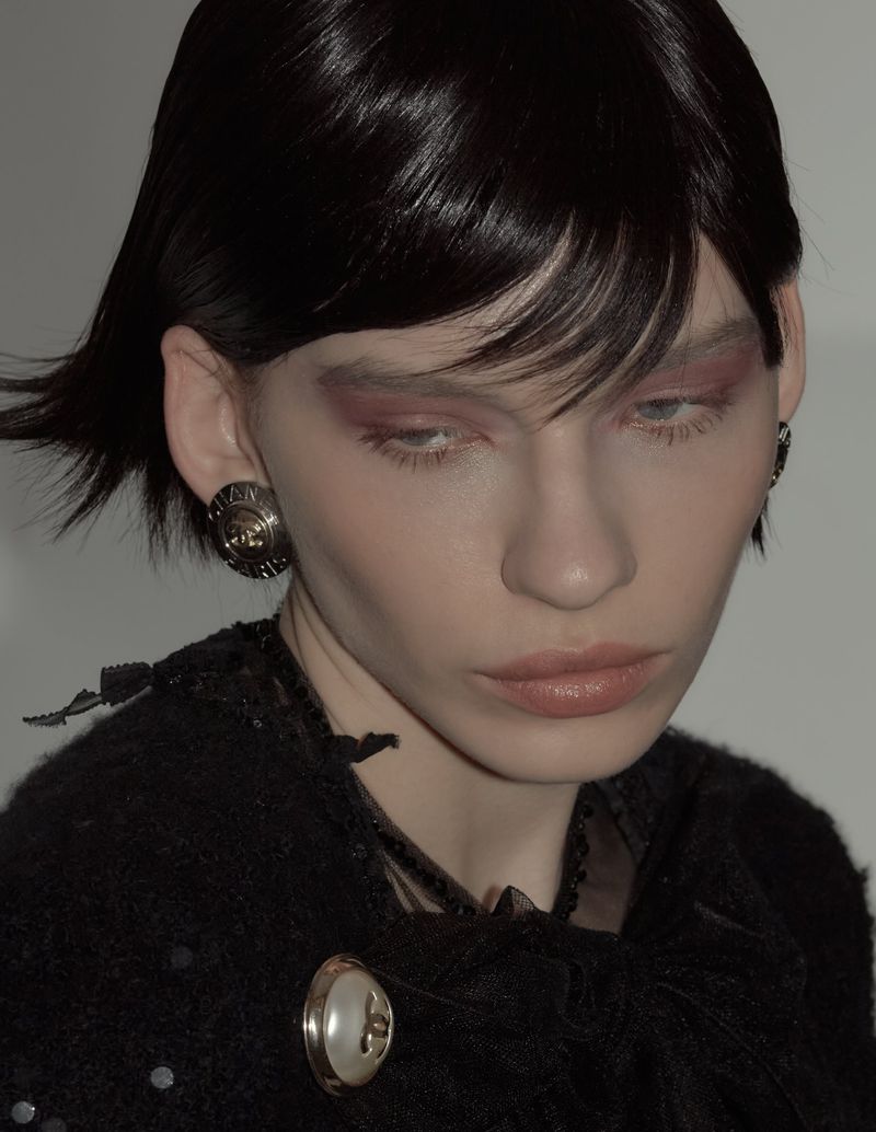 Close-up of a young woman with short dark hair, wearing a black tweed jacket with decorative buttons and earrings, indoors.
