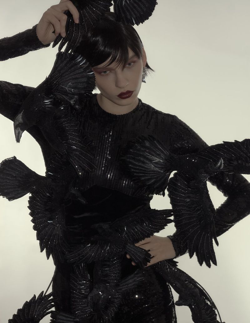 Fashion photo of a woman in a black, form-fitting dress adorned with black feathers, posing in front of a plain white background.