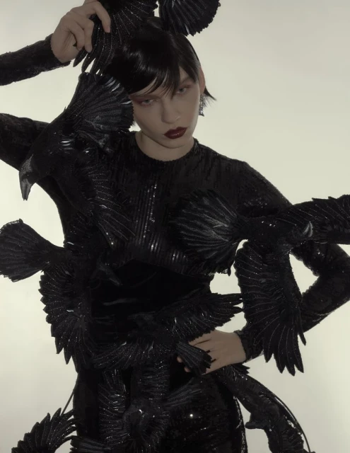 Fashion photo of a woman in a black, form-fitting dress adorned with black feathers, posing in front of a plain white background.