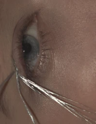 Extreme close-up of a model's blue eye with fine metallic silver wires resting across the lower eyelid and cheek.
