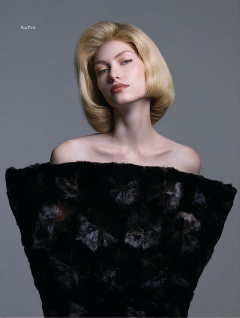 Elegant woman with blonde bob hairstyle and dark fur shawl poses against grey backdrop in a fashion portrait with soft lighting.