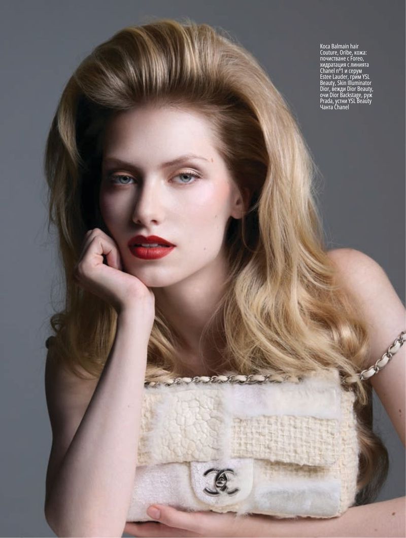 Blonde woman with red lipstick poses indoors with a textured Chanel handbag. Her hair is voluminous, and she rests her face on her hand.