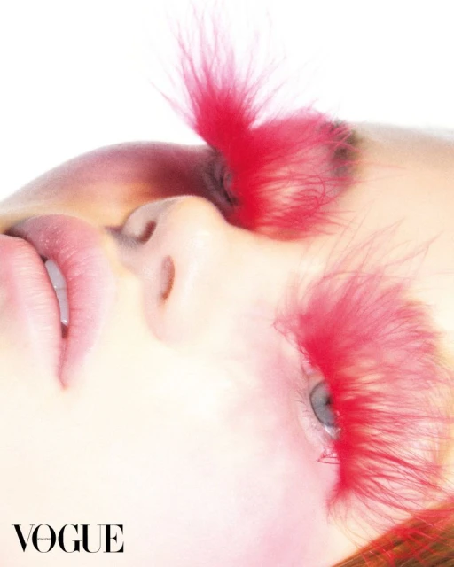 Close-up of a woman's face with pink feather eyelashes against a bright white background, creating a striking, fashion-forward aesthetic.