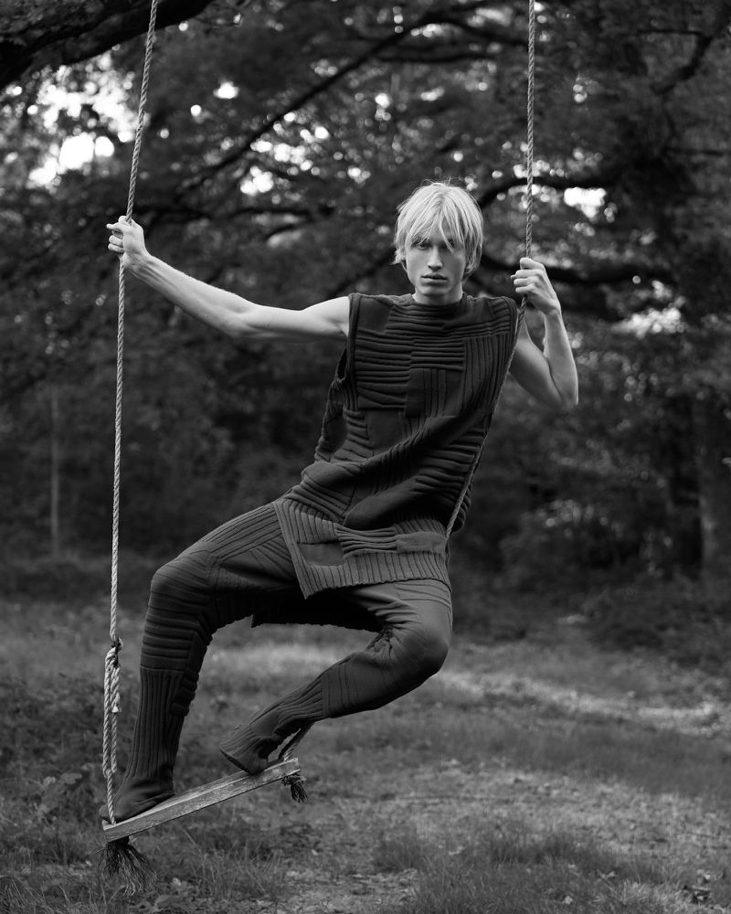 Monochrome fashion portrait of a male model in knitwear posing on a rope swing in a forest setting with dappled lighting.