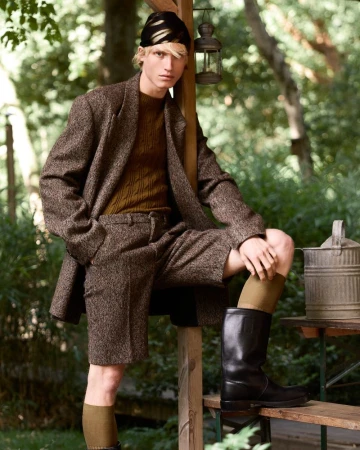 Male fashion model in tweed suit shorts and black boots poses outdoors in a garden setting with greenery and vintage lantern.