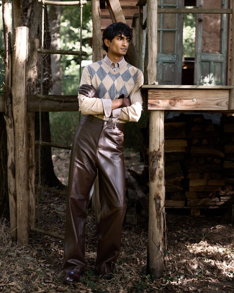 Adult male standing in front of a wood structure wearing leather pants, a collared shirt, and a sweater in an outdoor setting.