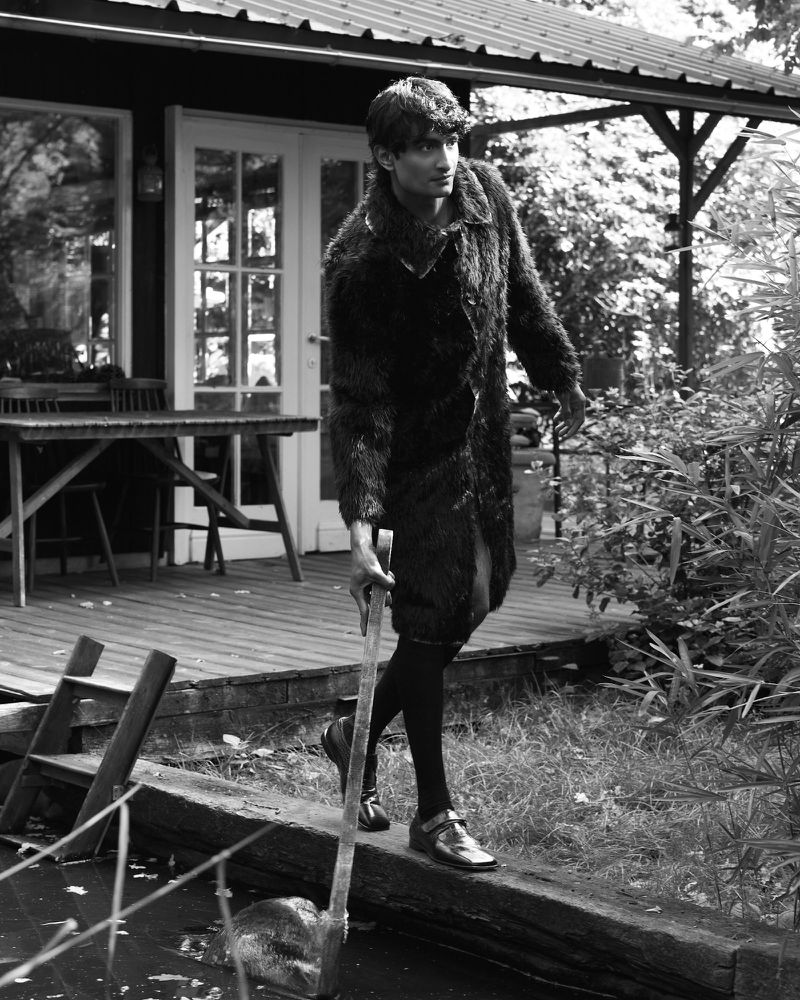 Black and white portrait of a person walking on a log near a body of water, wearing a fur coat, with a building and greenery in the background.