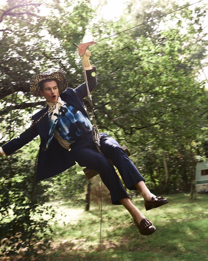 Adult male wearing a leopard print hat swinging in a wooded area with a trailer visible in the background on a sunny day.