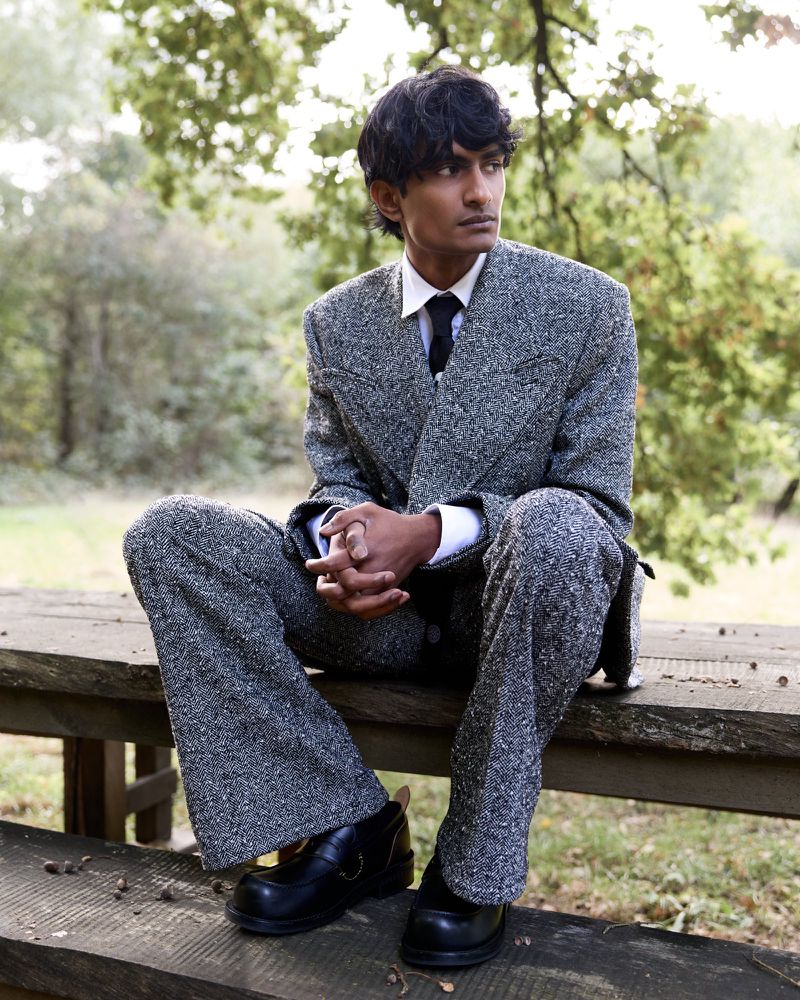 A person in a speckled gray suit sits on a wooden bench outdoors surrounded by trees in a medium shot, looking off to the side.