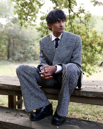 A person in a speckled gray suit sits on a wooden bench outdoors surrounded by trees in a medium shot, looking off to the side.